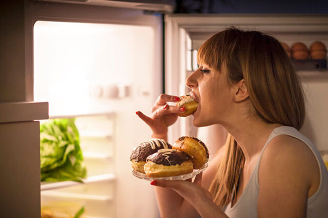 Foco na imagem de uma jovem mulher com transtorno alimentar fazendo um lanche da meia-noite, comendo rosquinhas na frente da geladeira